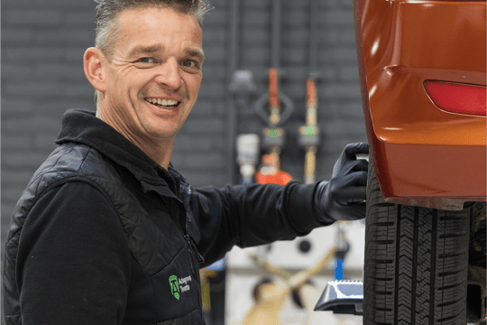 Een glimlachende medewerker van Autogroep Twente die in de werkplaats bezig is met een auto. Hij staat naast de auto en is bezig met het achterwiel.
