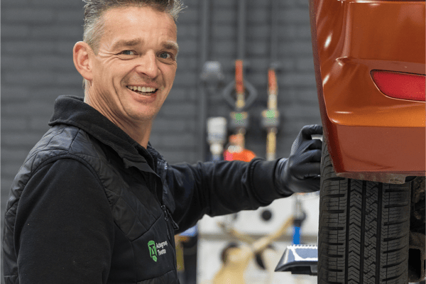 Een glimlachende medewerker van Autogroep Twente die in de werkplaats bezig is met een auto. Hij staat naast de auto en is bezig met het achterwiel.