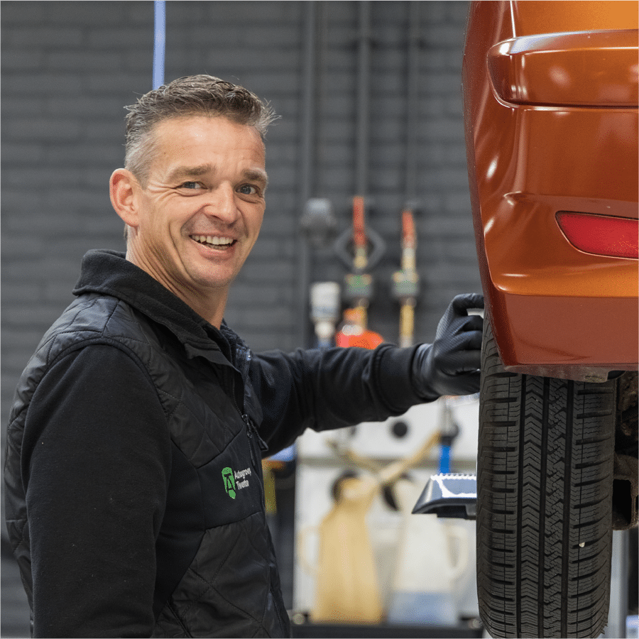 Een glimlachende medewerker van Autogroep Twente die in de werkplaats bezig is met een auto. Hij staat naast de auto en is bezig met het achterwiel.