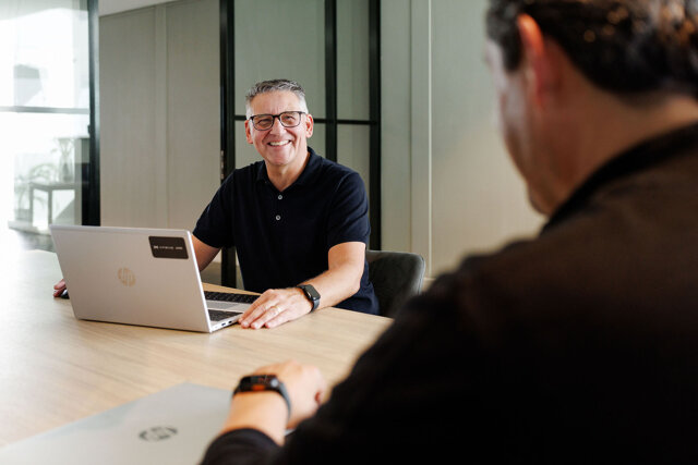 Medewerker werkt op een laptop aan een tafel, met een collega op de voorgrond die vraag stelt