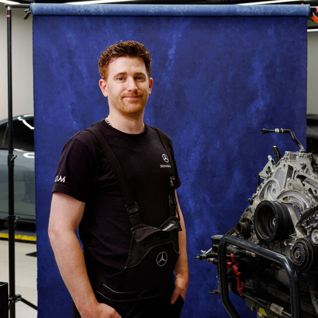 Jonge automonteur in zwart shirt staat voor een blauwe achtergrond naast een motorblok