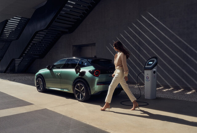 Vrouw loopt weg bij de laadpaal met daaraan de nieuwe Lancia Ypsilon Electric die aan het opladen is.